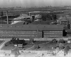 Le coton effectue un retour à Sherbrooke en 1918-1919, lorsque la compagnie <em>Canadian Connecticut Cotton Mills</em> y construit une usine, rue du Pacifique. Elle y fabrique des toiles de pneu en coton. L’usine est rachetée en 1928 par la <em>Dominion Textile Co.</em> La compagnie est marquée par plusieurs grèves entre 1936 et 1938; l’attitude patronale envers ses employés engendre d’ailleurs le déclenchement d’une Commission royale d’enquête sur l’industrie du textile en 1936. Les usines de Sherbrooke ferment progressivement entre 1994 et 2002. Photo : L’usine de la <em>Dominion Textile</em>, installée entre la voie ferrée et le quartier ouvrier (1937).