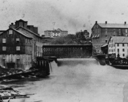 Le barrage no 5 et le confluent des rivières Magog et Saint-François, vers 1885. Le barrage alimente en eau les moulins logés dans les bâtiments situés sur les deux rives. L’eau est alors la principale source d’énergie pour les manufactures de la ville, ce pour quoi celles-ci sont concentrées le long de la rivière Magog.