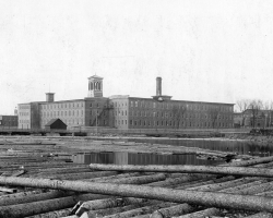 La rivière Magog recouverte de billots de bois, destinés à la scierie de la <em>British American Land Company</em>, en 1898. À l’arrière, les bâtiments de la Paton.