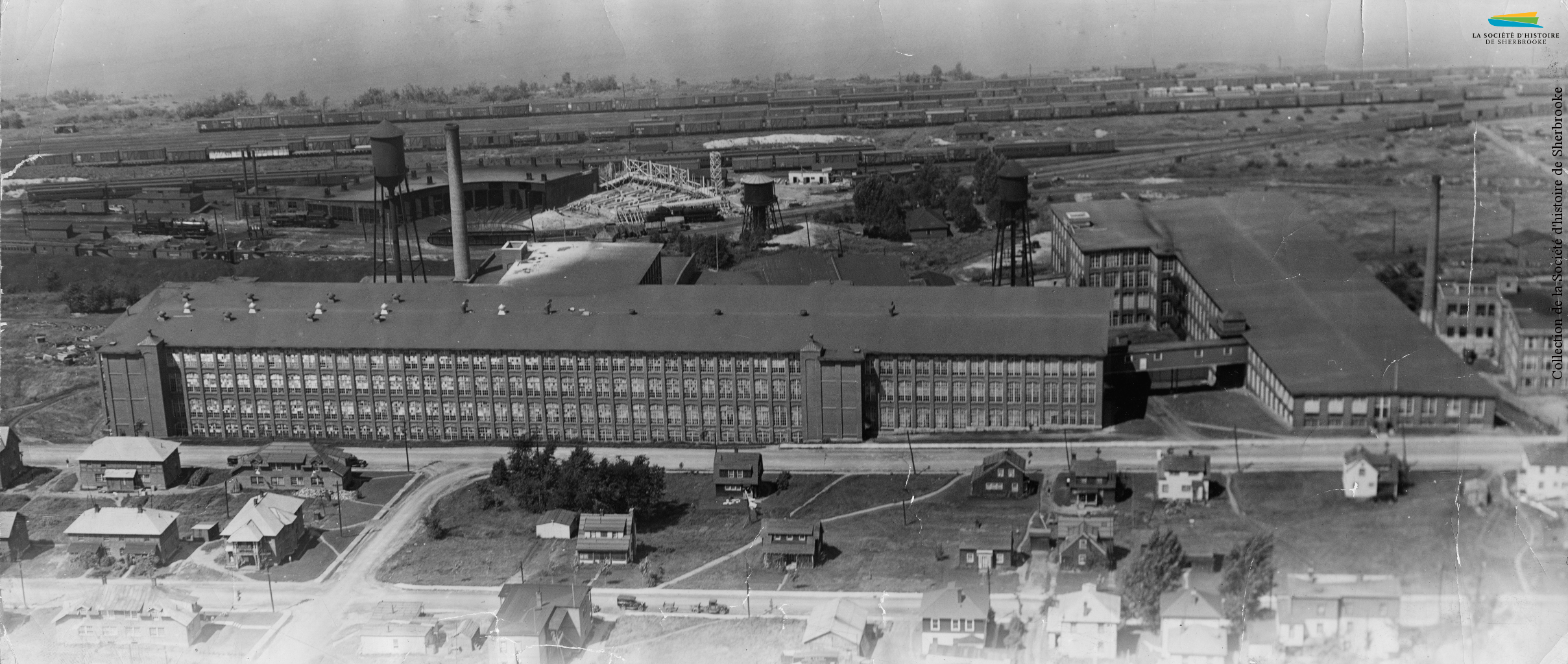 Le coton effectue un retour à Sherbrooke en 1918-1919, lorsque la compagnie <em>Canadian Connecticut Cotton Mills</em> y construit une usine, rue du Pacifique. Elle y fabrique des toiles de pneu en coton. L’usine est rachetée en 1928 par la <em>Dominion Textile Co.</em> La compagnie est marquée par plusieurs grèves entre 1936 et 1938; l’attitude patronale envers ses employés engendre d’ailleurs le déclenchement d’une Commission royale d’enquête sur l’industrie du textile en 1936. Les usines de Sherbrooke ferment progressivement entre 1994 et 2002. Photo : L’usine de la <em>Dominion Textile</em>, installée entre la voie ferrée et le quartier ouvrier (1937).