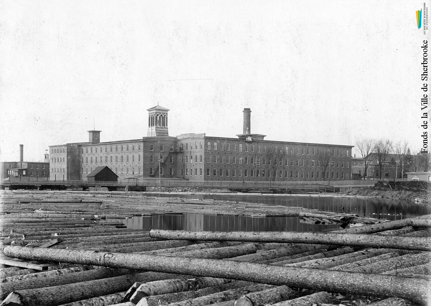 La rivière Magog recouverte de billots de bois, destinés à la scierie de la <em>British American Land Company</em>, en 1898. À l’arrière, les bâtiments de la Paton.